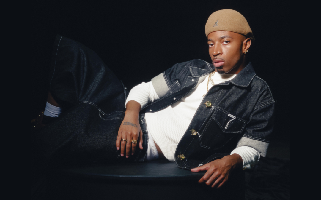 Tyrik J Patterson poses for a photo, lounging on one side with one leg crossed over the other. He wears dark jeans, a tan hat, and a short-sleeve denim shirt opened to reveal another white shirt underneath.