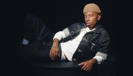 Tyrik J Patterson poses for a photo, lounging on one side with one leg crossed over the other. He wears dark jeans, a tan hat, and a short-sleeve denim shirt opened to reveal another white shirt underneath.