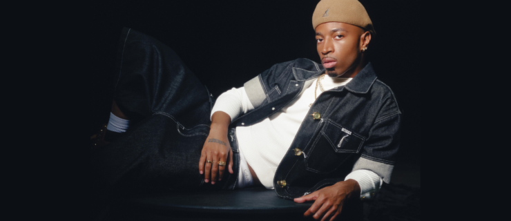 Tyrik J Patterson poses for a photo, lounging on one side with one leg crossed over the other. He wears dark jeans, a tan hat, and a short-sleeve denim shirt opened to reveal another white shirt underneath.