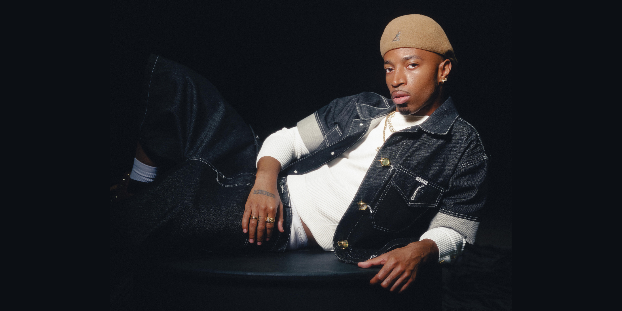 Tyrik J Patterson poses for a photo, lounging on one side with one leg crossed over the other. He wears dark jeans, a tan hat, and a short-sleeve denim shirt opened to reveal another white shirt underneath.