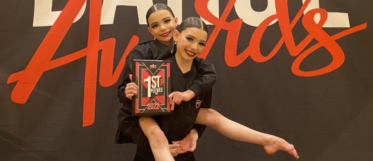 Sylvie Win and Ellary Day Szyndlar pose for a photo together in front of a The Dance Awards step and repeat. Ellary Day holds Sylvie Win on her back, and Sylvie Win shows off their first place Peewee Division plaque.
