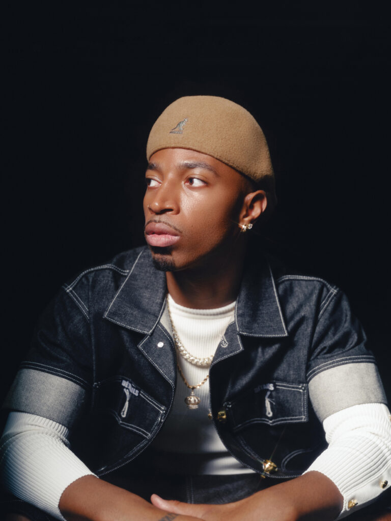 Tyrik J Patterson poses for a portrait, resting his arms in front and crossing his hands casually as he looks off to the side. He wears a tan hat and a short-sleeve denim shirt opened to reveal another white shirt underneath.