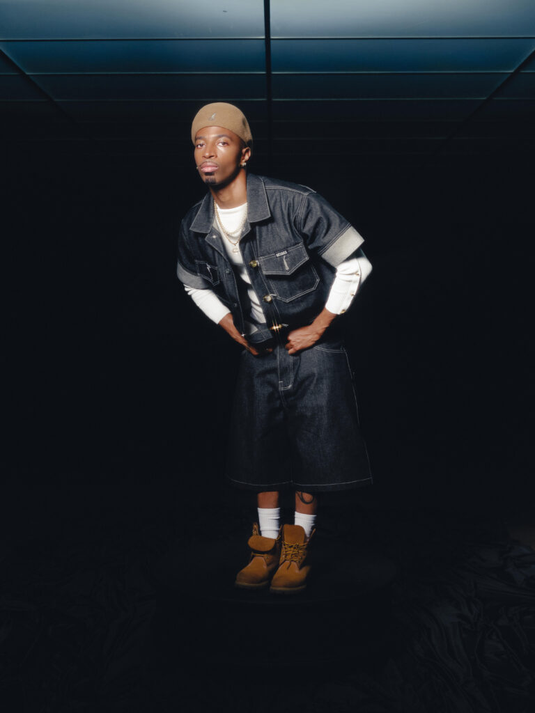 Tyrik J Patterson wears dark jeans, a tan hat, light brown boots, and a short-sleeve denim shirt opened to reveal another white shirt underneath. He stands with his legs together, his hands in his pockets as he leans his upper body forward and slightly to the side.