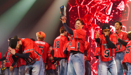 On a dramatically lit stage, a crowd of dancers in red jerseys celebrates. At the front of the group, one dancer grins broadly as he hoists a black trophy.