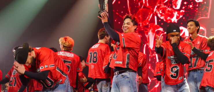 On a dramatically lit stage, a crowd of dancers in red jerseys celebrates. At the front of the group, one dancer grins broadly as he hoists a black trophy.