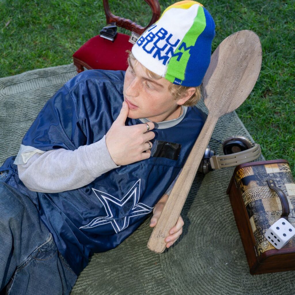 Dossan Bell leans on one elbow while resting on a blanket on the grass. He holds a large wooden paddle in one hand, the other lifted to rest his thumb on his lower lip as he looks away. He wears a blue jersey and a beanie that says "Bum bum bum bum."
