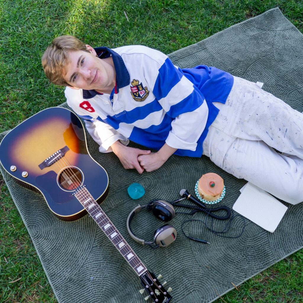 Sean Garrity smizes at the camera as he leans on one side, one hand resting on the other. He lies on a blanket on the grass with a guitar, headphones, and a donut arranged nearby.