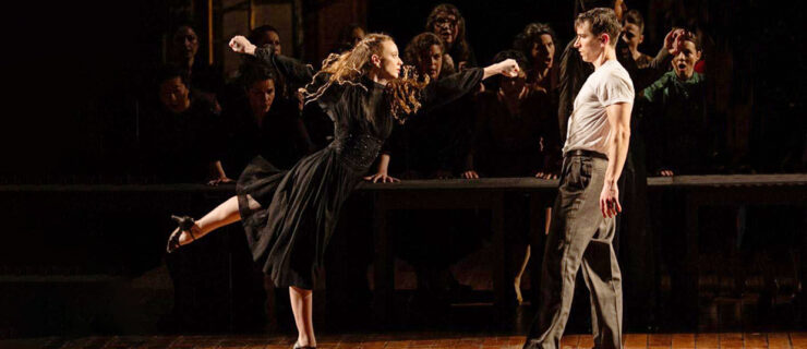 Payton Johnson performs an arabesque while reaching towards Alexander Bozinoff. She wears a long sleeve, knee-length black dress and character heels.