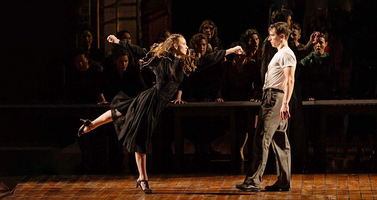 Payton Johnson performs an arabesque while reaching towards Alexander Bozinoff. She wears a long sleeve, knee-length black dress and character heels.