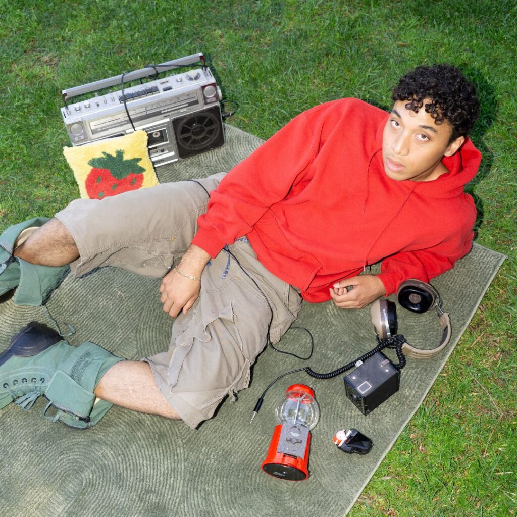James Herron rests on one elbow while lounging on a blanket on the grass. He looks up at the camera and wears a bright red hoodie. Around him are miscellaneous objects like a boombox and bubblegum dispenser.