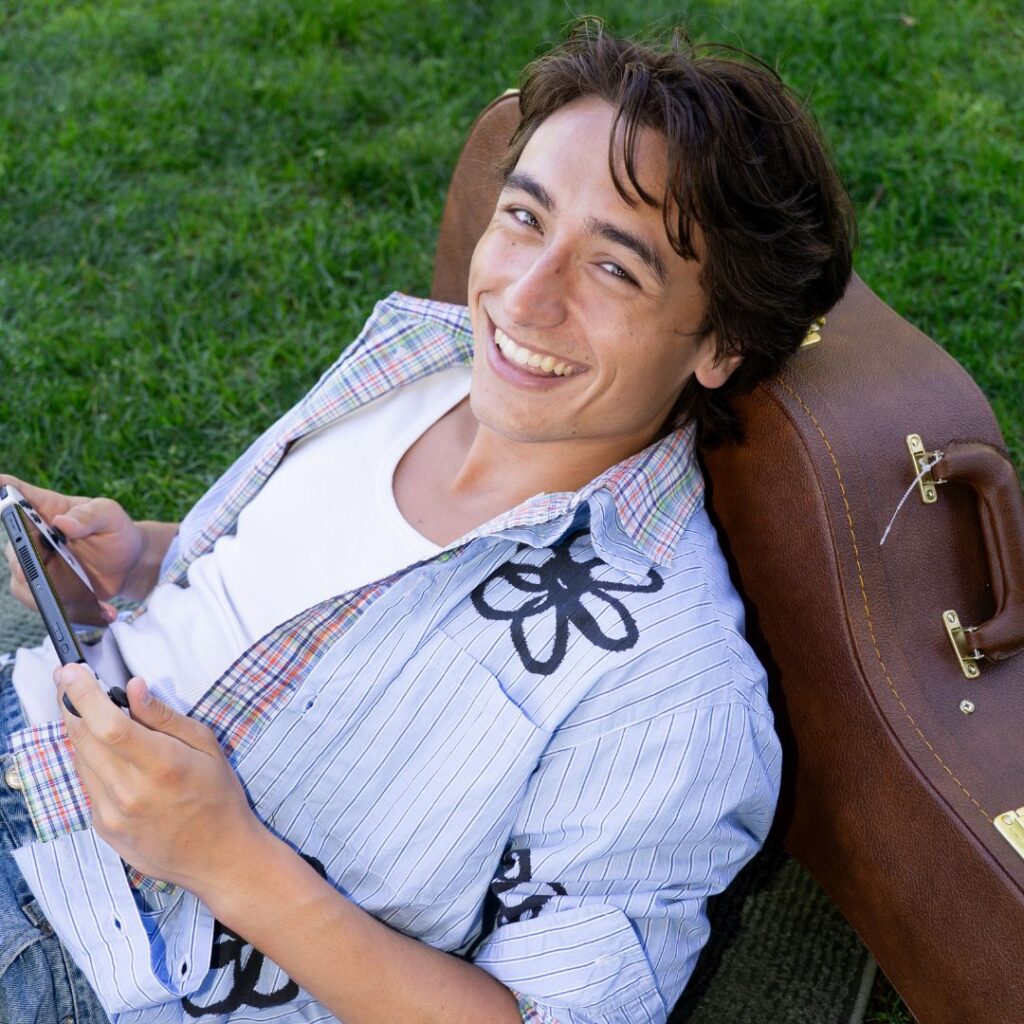 Jagger Ray Moon, in a button down with flowers, leans back on a guitar case while lounging on a blanket on the grass. He smiles at the camera while holding a phone horizontally.