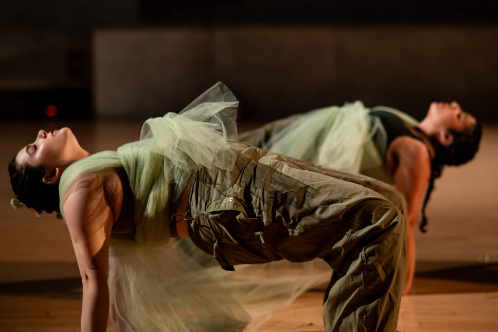 Two dancers in light green costumes with tulle pose in bridge positions, with their upper bodies and thighs parallel to the floor, arms and lower legs perpendicular for support. They close their eyes with their faces upward. One dancer is positioned in front of the other.