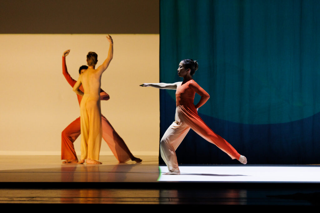 Onstage in front of a background with light yellow on the left and dark blue on the right, three dancers perform wearing orange, yellow, and red flowy pantsuits. In front, one dancer lunges forward with one arm tucked behind and the other extended in front. Behind, two dancers face each other—one lunging sideways and the other standing in parallel—each with one arm lifted.