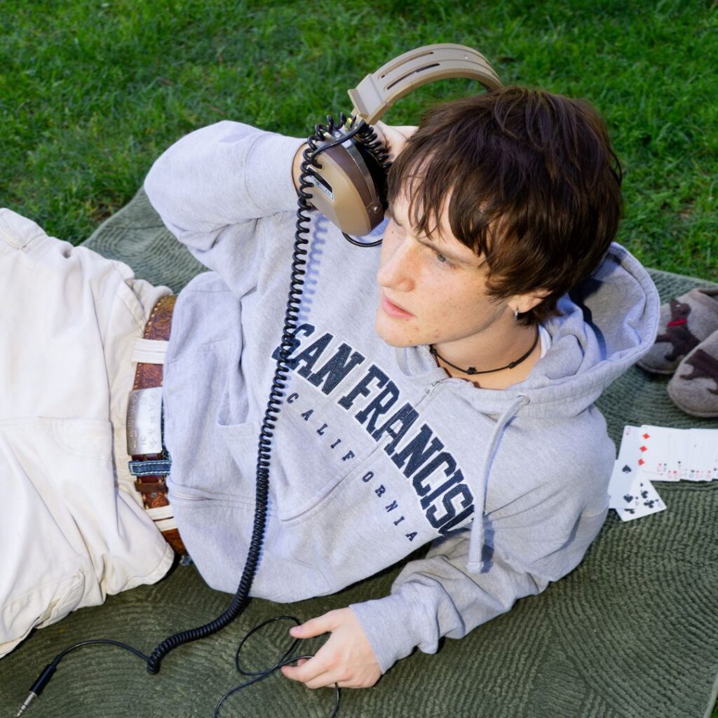 Ollie Hincy looks offscreen as he lounges on a blanket on the grass, holding a pair of headphones up to one ear. He wears a San Francisco sweatshirt, and a keyboard rests nearby.