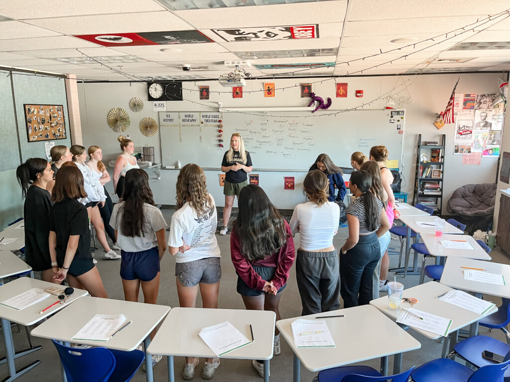 In a high school classroom, a group of dance team members stand in the middle of a large circle of desks. Dr. Chelsea Pierotti addresses them in front of a whiteboard.