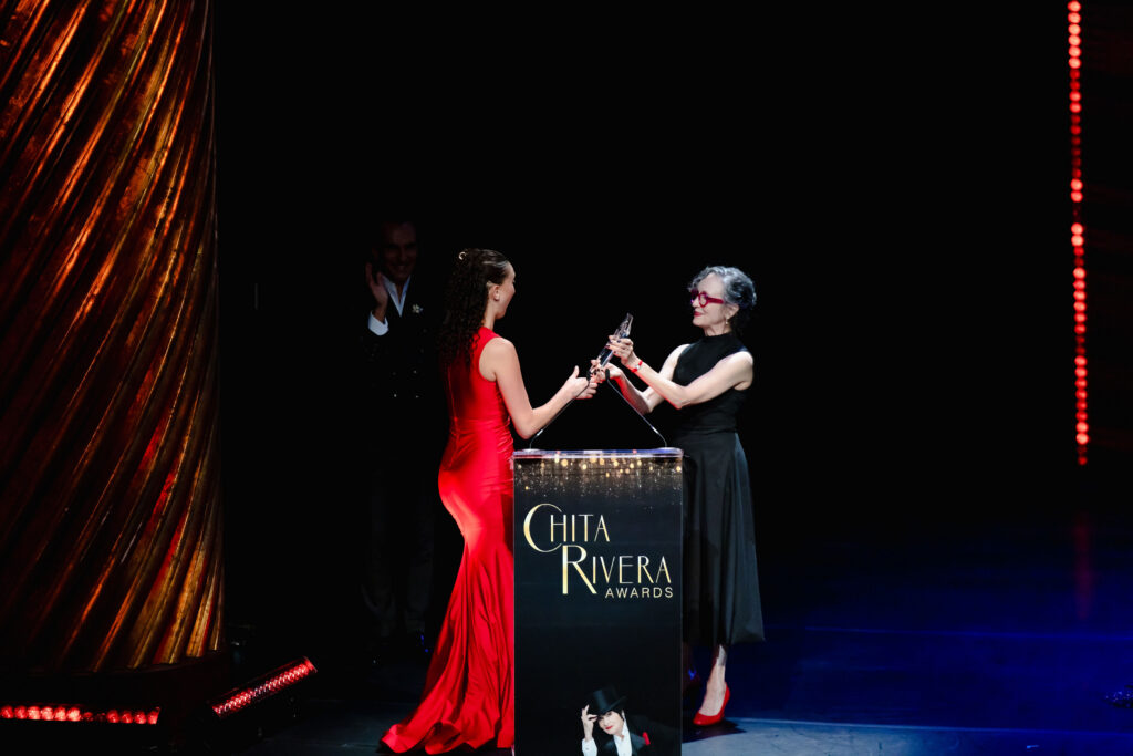 Helena Padial, in a red evening gown, accepts a glass award from Bebe Neuwirth, who wears a black dress with red glasses and red heels. In front of them is a podium with the Chita Rivera Awards logo and a photo of Rivera.