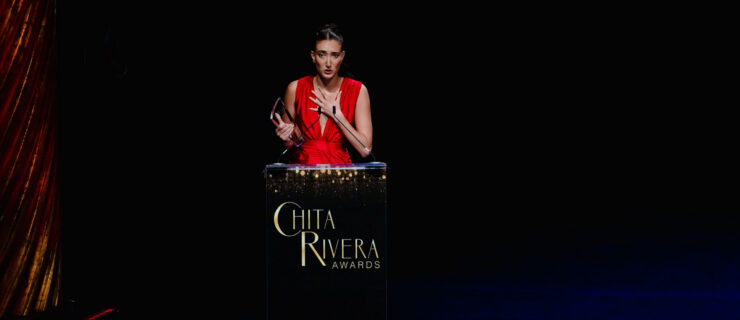 Helena Padial, in a red gown, holds a glass award as she gives an acceptance speech. She stands behind a podium with the Chita Rivera Awards logo.