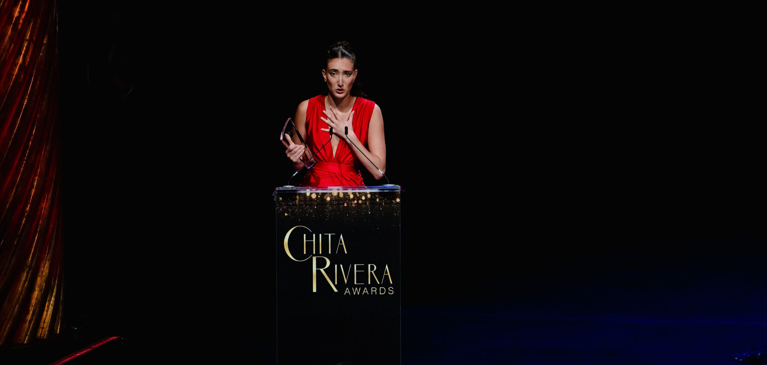 Helena Padial, in a red gown, holds a glass award as she gives an acceptance speech. She stands behind a podium with the Chita Rivera Awards logo.