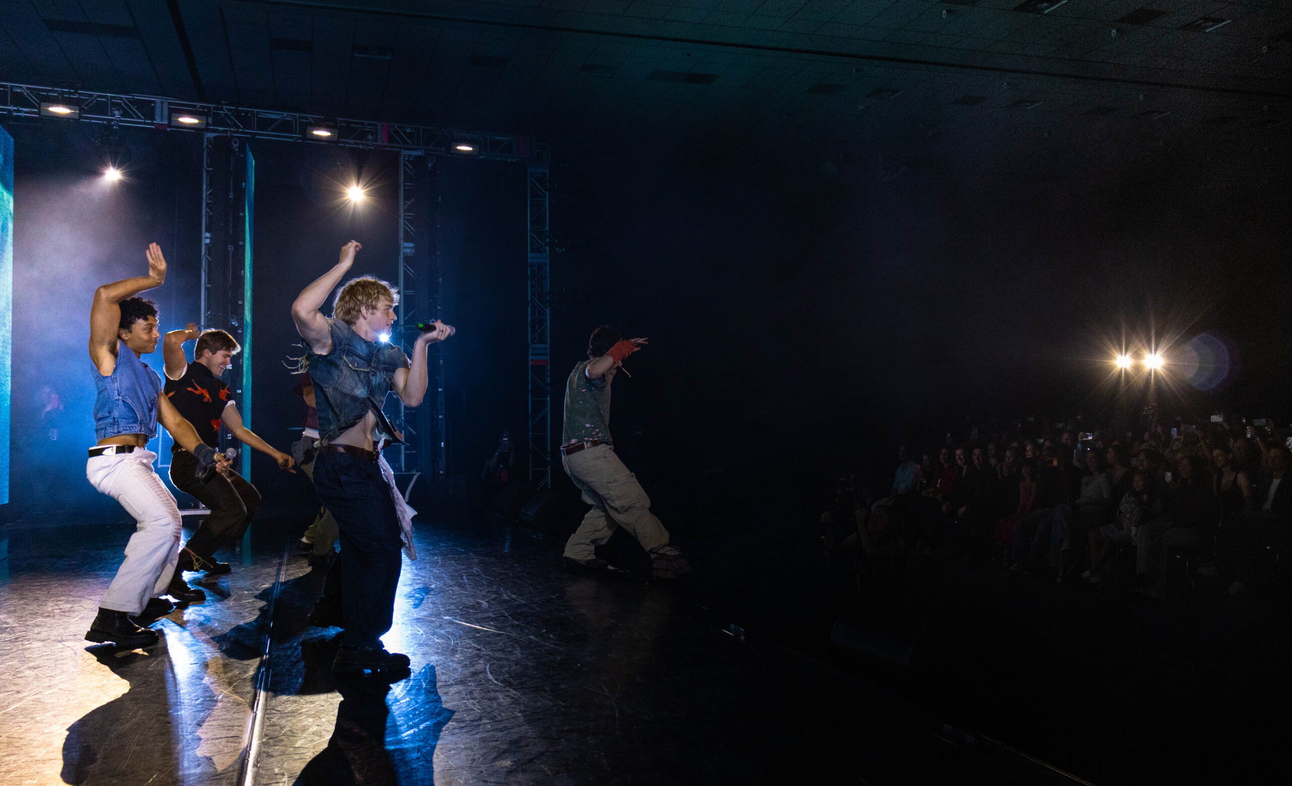 Onstage at a dance convention, the Full Circle Boys perform together, singing and dancing in synchrony. They wear various colors and styles of jeans and denim tops.
