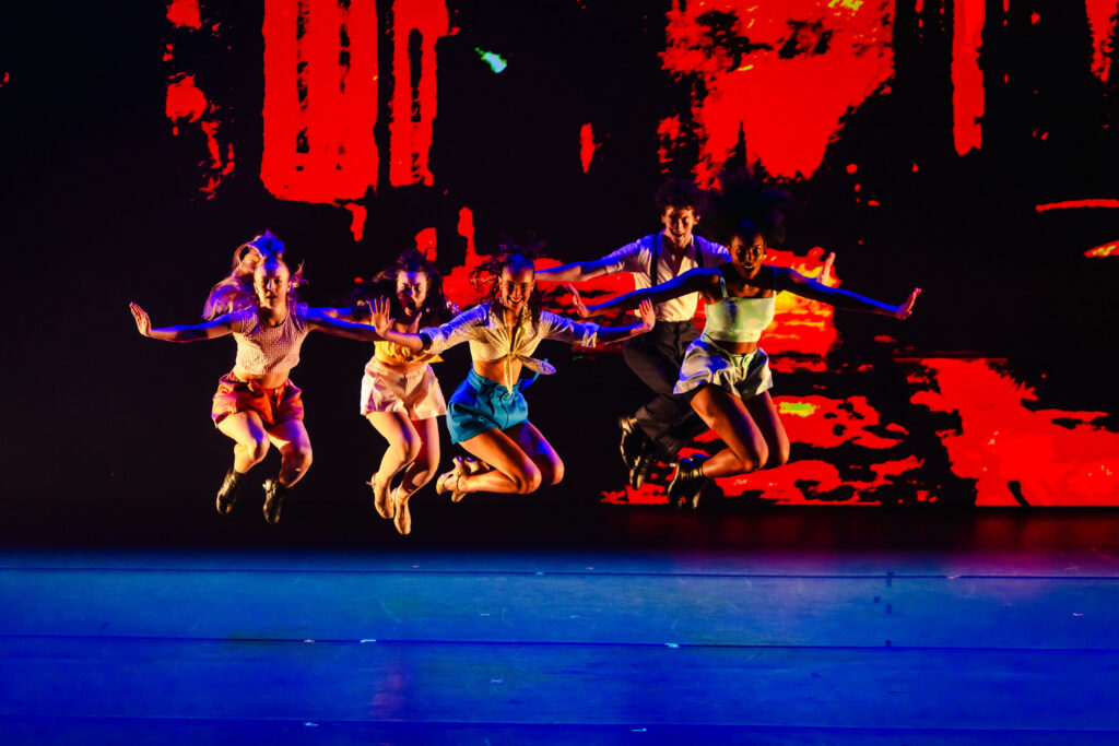 Five performers jump together onstage with their legs tucked and arms extended outward, hands flexed.