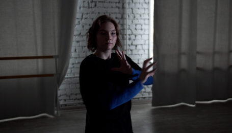 A young dancer in a dark blue long-sleeved shirt dances in a darkly lit studio. She bents her elbows and moves her arms inward, fingers separated with slight curves.