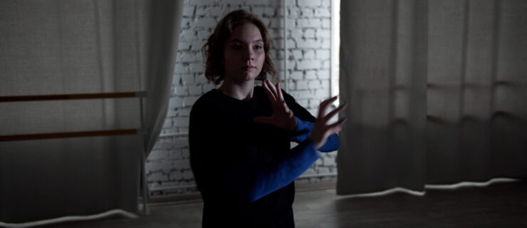 A young dancer in a dark blue long-sleeved shirt dances in a darkly lit studio. She bents her elbows and moves her arms inward, fingers separated with slight curves.