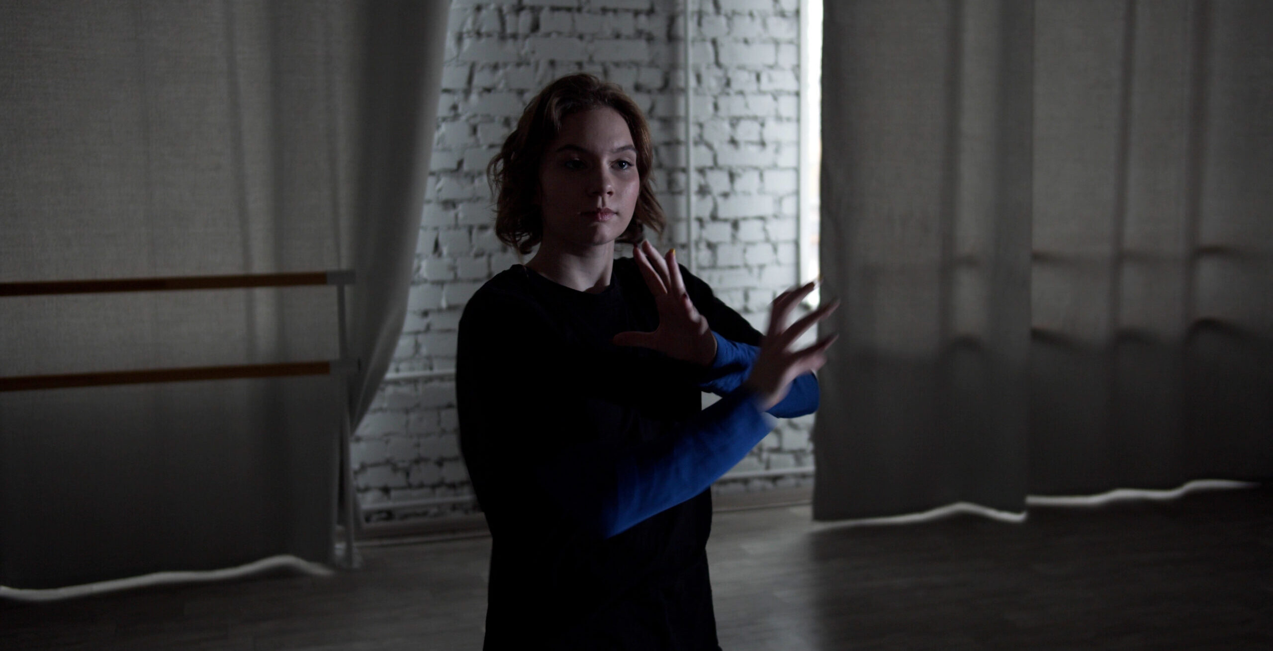 A young dancer in a dark blue long-sleeved shirt dances in a darkly lit studio. She bents her elbows and moves her arms inward, fingers separated with slight curves.