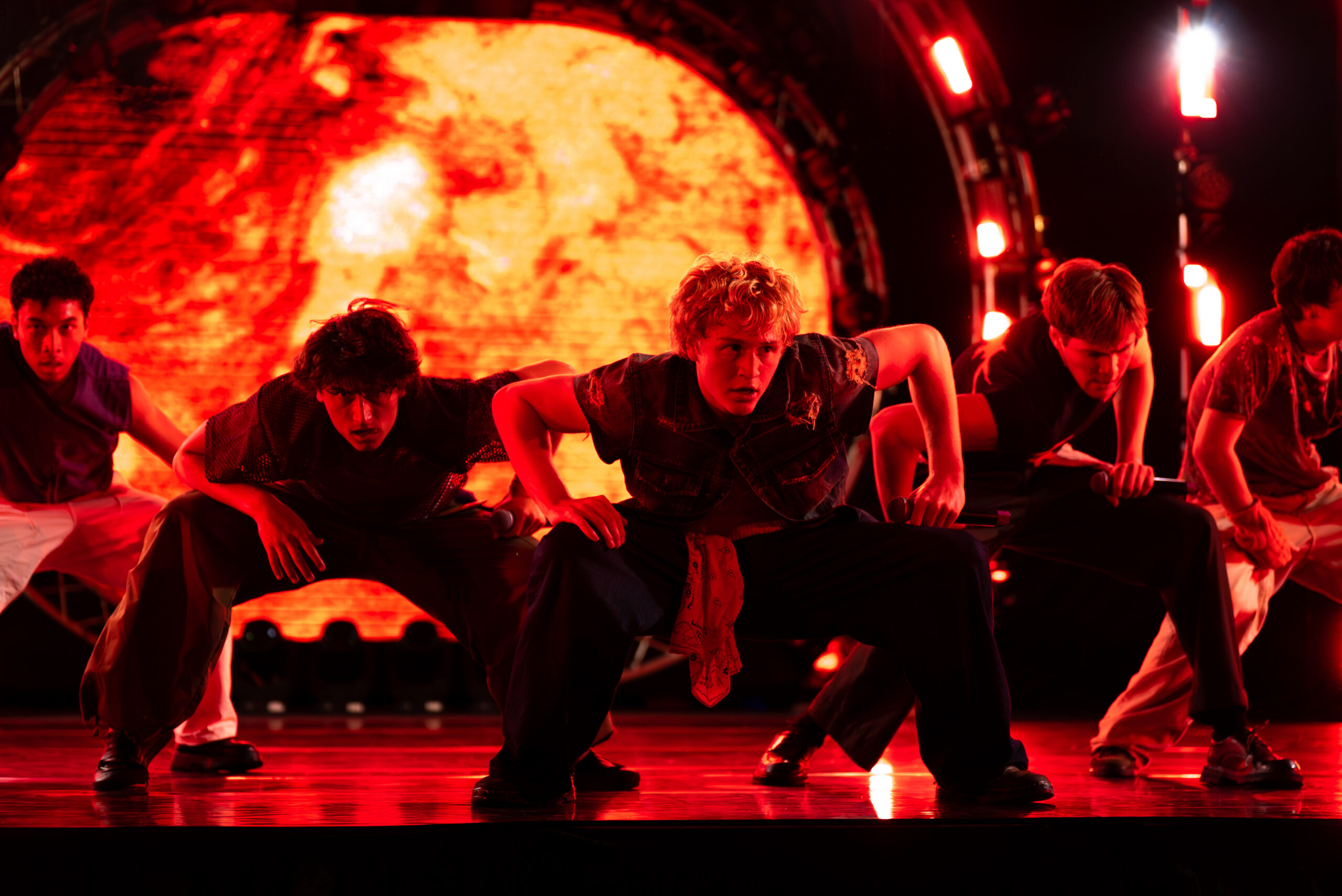 In dark red lighting in front of a fiery projection, the Full Circle Boys dance together onstage. They bend both legs and lean forward, hands or arms on their thighs, with a slight twist in their upper bodies.