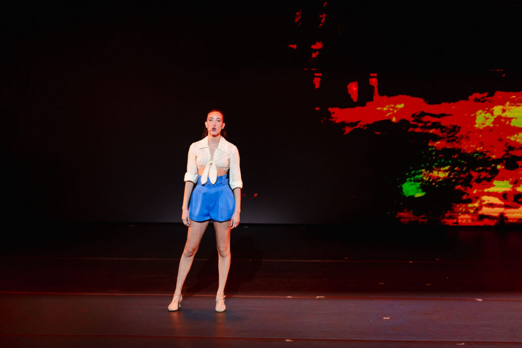 Helena Padial, in a white cropped shirt and blue shorts, stands onstage and sings.