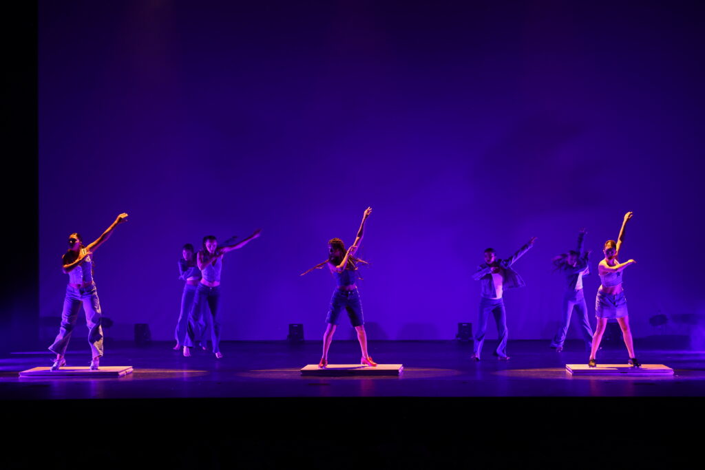 Onstage in dark purplish lighting, seven dancers perform, with three in the front on separate tap boards. They swing their arms up to the left while moving their feet.
