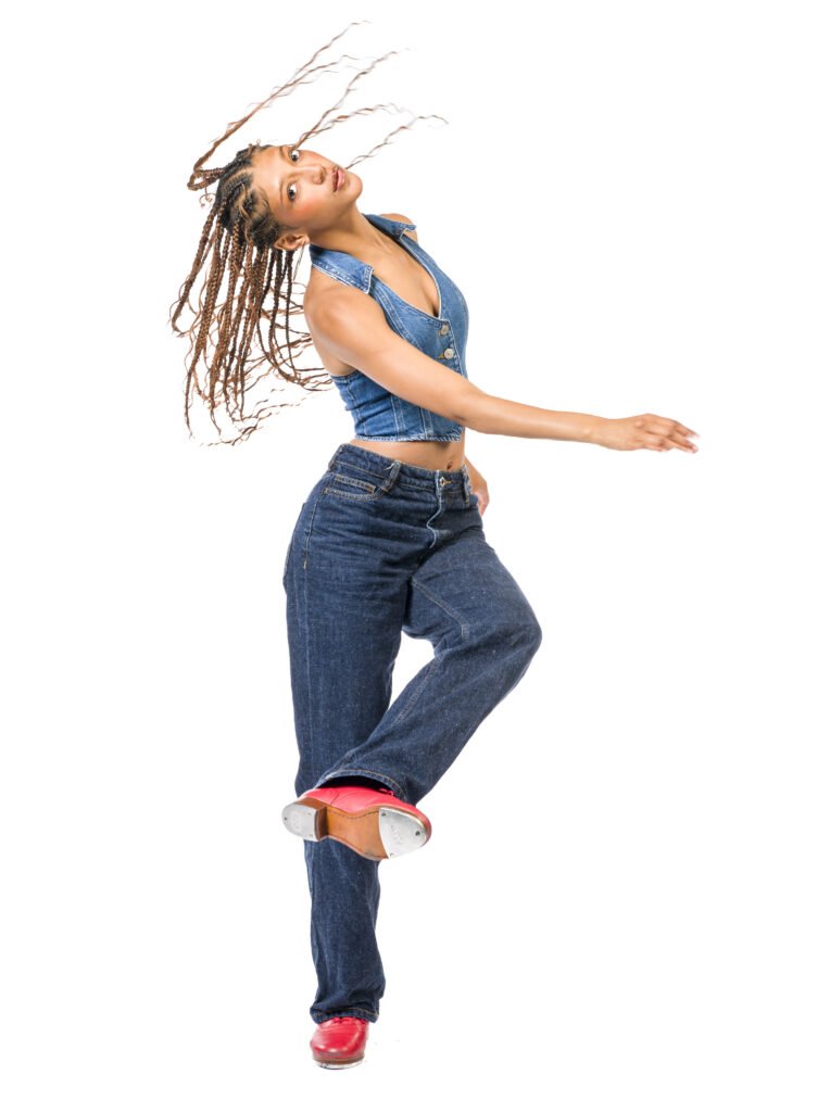 In front of a bright white background, Rio Gomez lifts and crosses one leg in front, showing the taps on the bottom of her shoe, while twisting her torso with one arm across. Her long braids fly. She wears jeans, a denim halter top, and red tap shoes.
