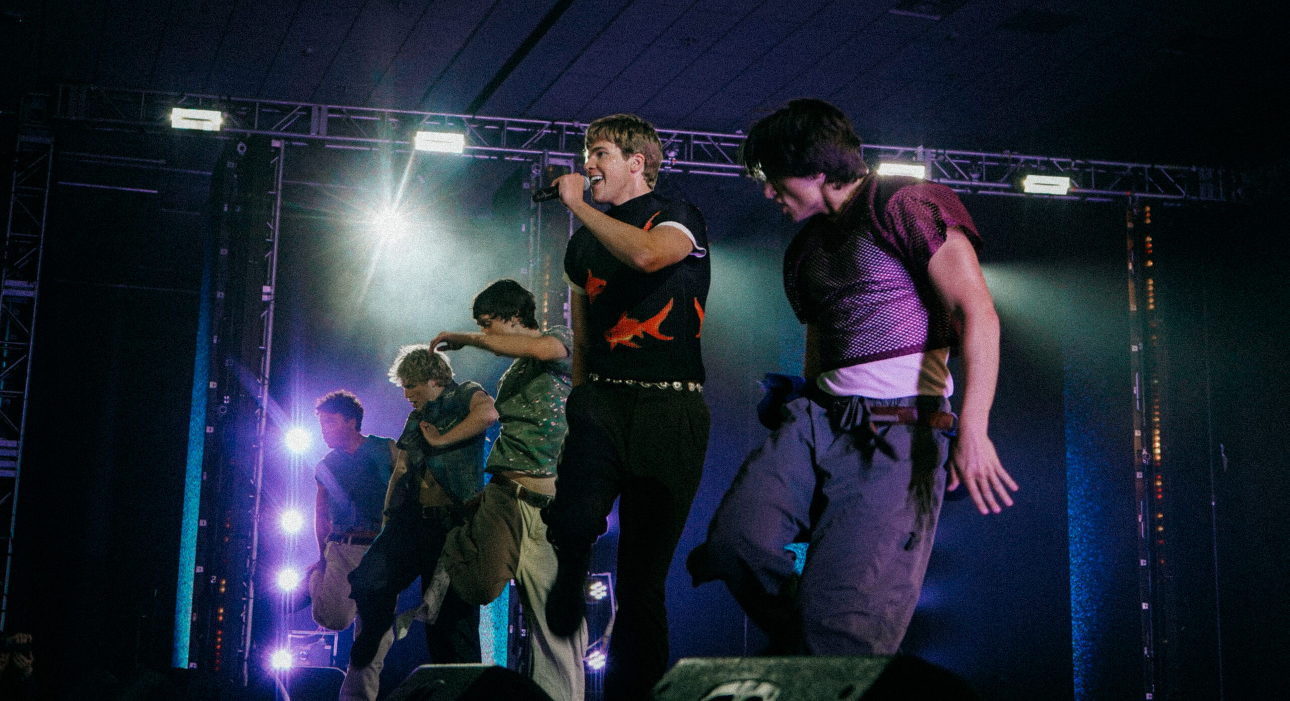 The Full Circle Boys perform together onstage, lifting one leg up with hips twisting to the side. One of the boys sings into a microphone.