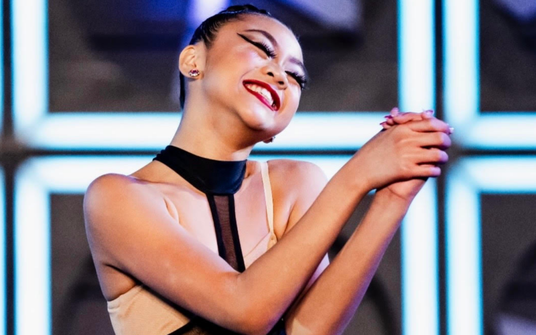 A close-up of Madisyn Rose Amos, in costume onstage, smiling widely with her hands clasped and arms bent in a show of gratitude.
