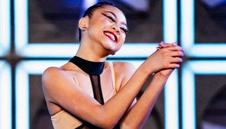 A close-up of Madisyn Rose Amos, in costume onstage, smiling widely with her hands clasped and arms bent in a show of gratitude.