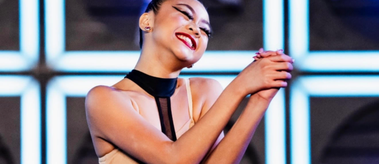 A close-up of Madisyn Rose Amos, in costume onstage, smiling widely with her hands clasped and arms bent in a show of gratitude.
