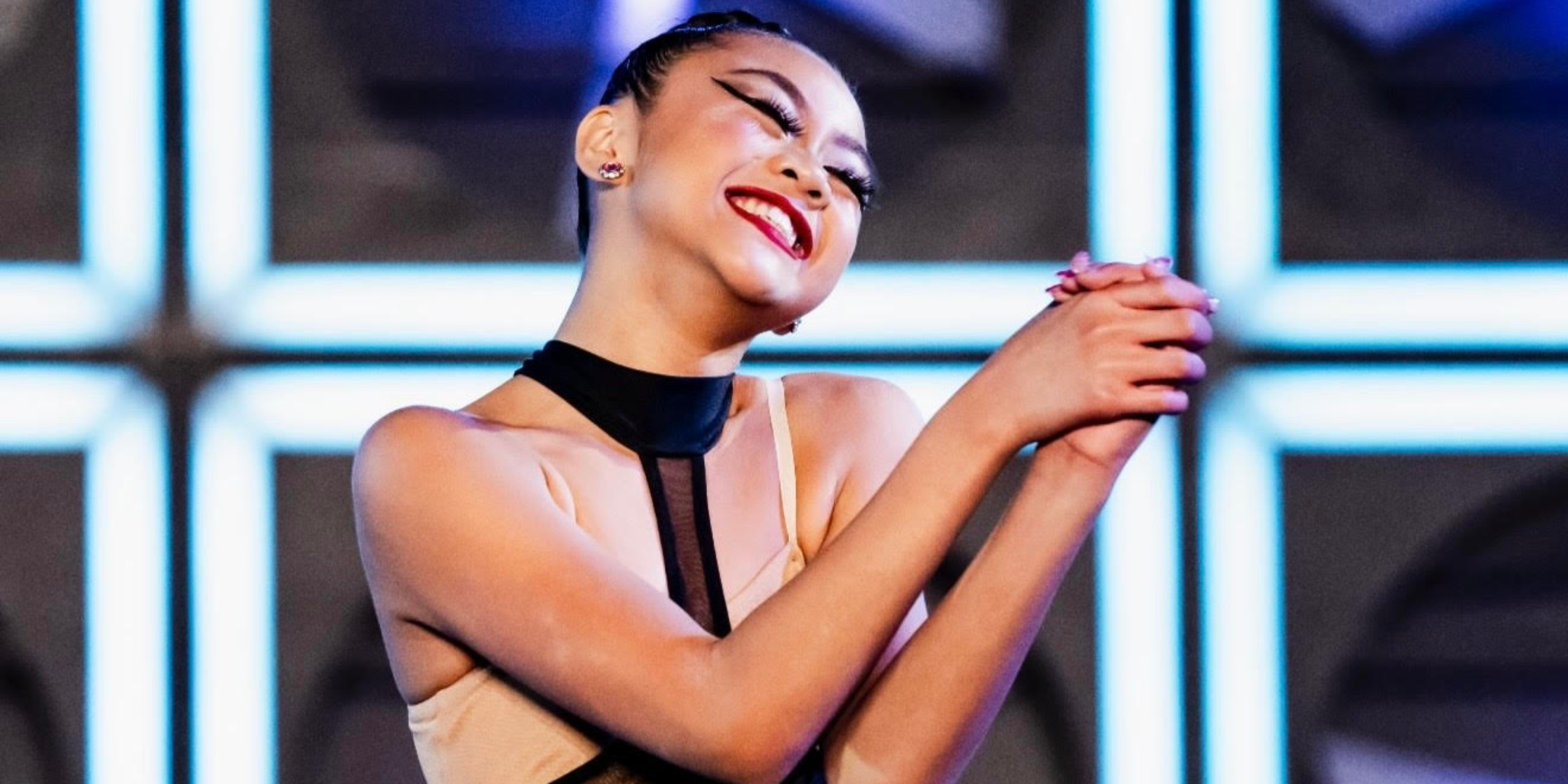 A close-up of Madisyn Rose Amos, in costume onstage, smiling widely with her hands clasped and arms bent in a show of gratitude.