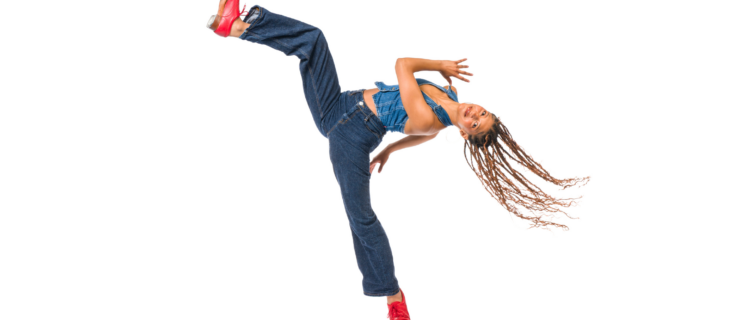 In front of a bright white background, Rio Gomez pitches back with one leg lifted up in the air, her hair flying. She pumps her arms and looks toward the camera with a smile. She wears jeans, a denim halter top, and red tap shoes.