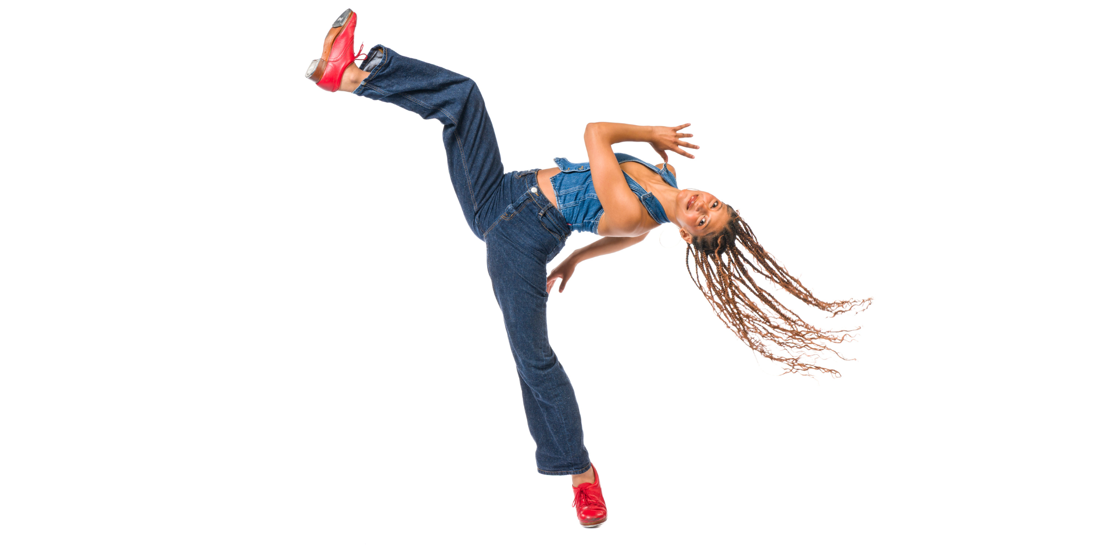 In front of a bright white background, Rio Gomez pitches back with one leg lifted up in the air, her hair flying. She pumps her arms and looks toward the camera with a smile. She wears jeans, a denim halter top, and red tap shoes.