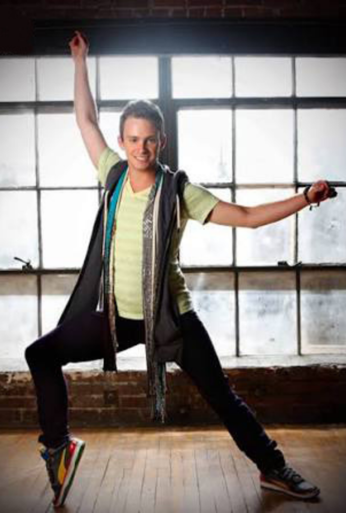 Jakob Karr lunges to the side with his arms raised, wearing jeans, a light green shirt, and a gray vest. He dances in front of a warehouse window.