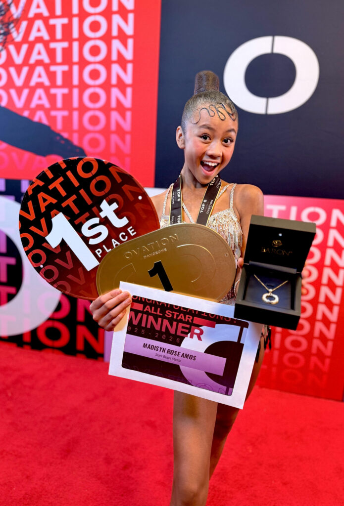Madisyn Rose Amos, with a slick ballroom hairstyle and wearing a shimmery costume, poses in front of an Ovation Dance wall while holding several awards.