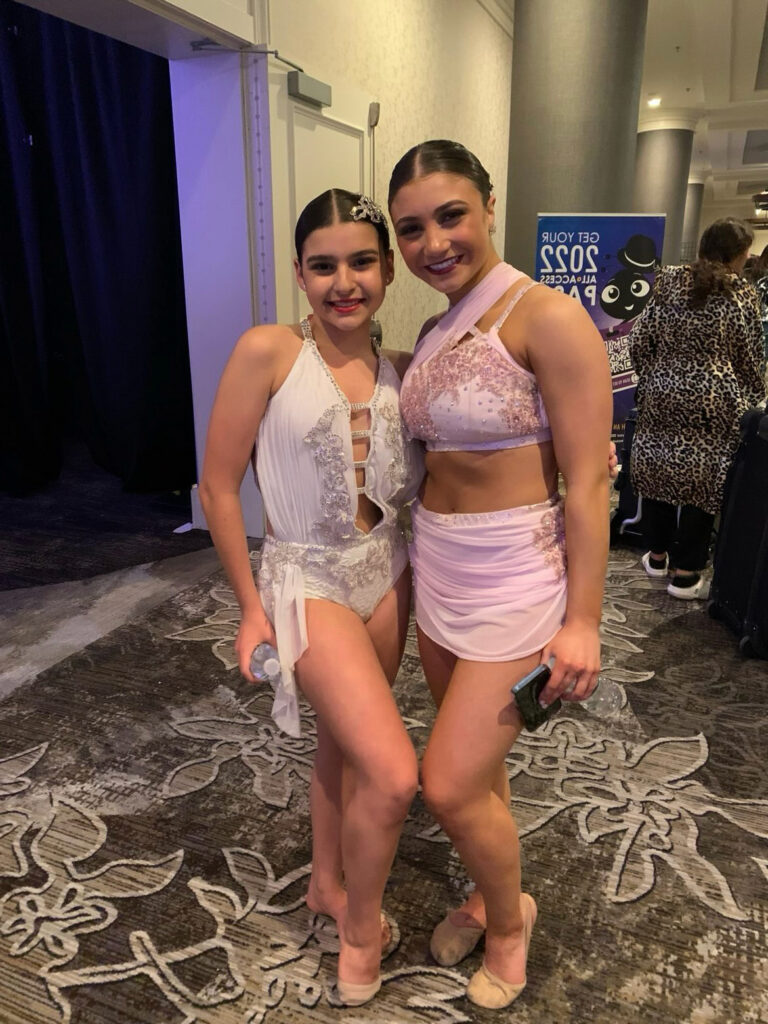 Two dancers pose together in a hallway at a dance competition. They wear pink and white costumes with short skirts and ornate tops.