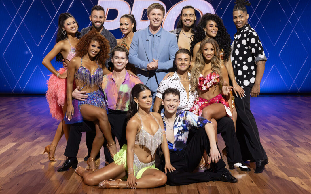A group of dancers in various ballroom and Latin dance costumes pose for a group photo surrounding Robert Irwin, who wears a powder-blue suit.