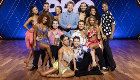A group of dancers in various ballroom and Latin dance costumes pose for a group photo surrounding Robert Irwin, who wears a powder-blue suit.