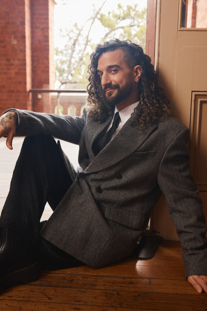 Mark Ballas, in an oversized gray suit, sits casually in a doorway with one leg tucked up and one arm resting on his knee. He smizes at the camera and wears his long, curly hair down.