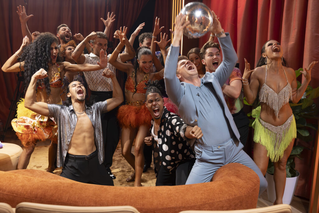 A group of dancers in costume gather behind Robert Irwin as he lifts a disco ball. They all cheer and lift their arms.