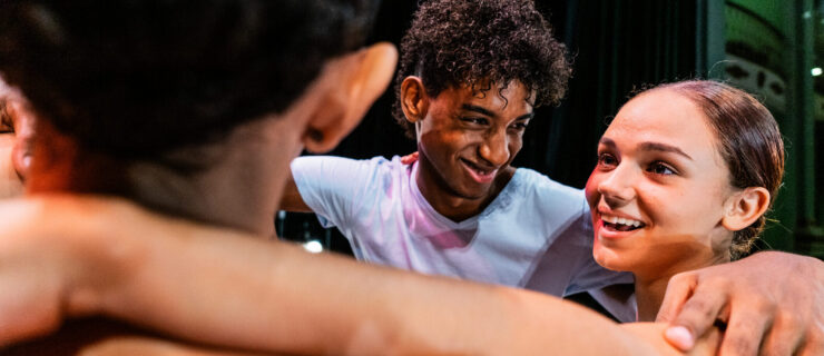 A group of teenage dancers huddle for a group hug onstage.