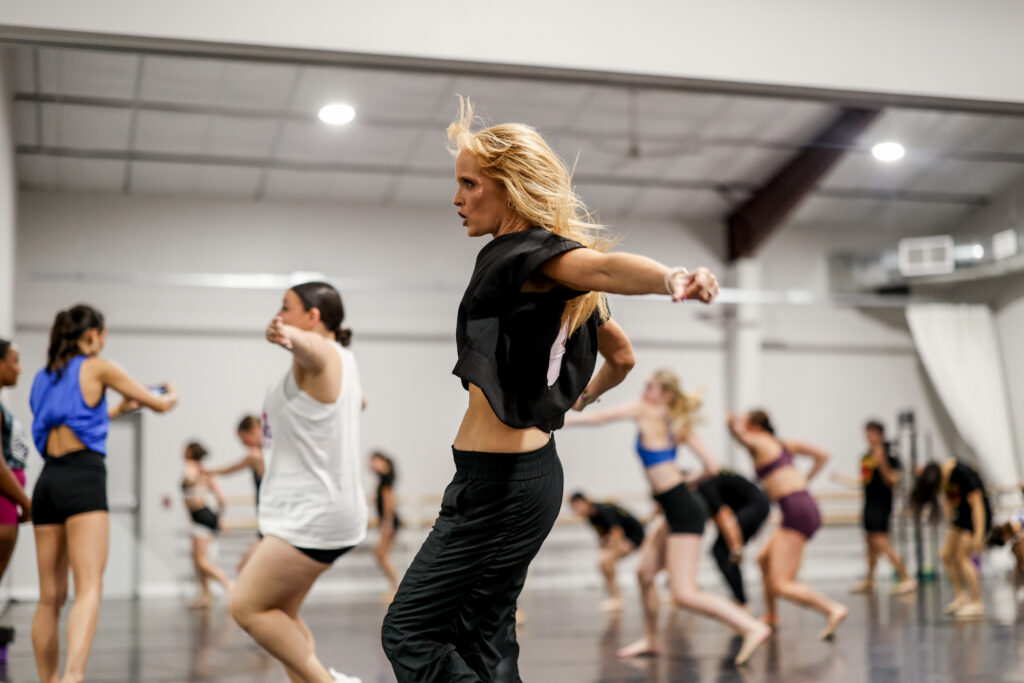 Michelle Leagans, in a flowy black crop top and black pants, leads a dance class in a large studio. Students mimic her movement behind her as she steps forward and pumps one arm out to the side.