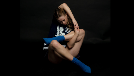 In front of a pitch-black backdrop, Lucy Turner sits with her limbs bent up and crossed in a pretzel-like shape, one arm reaching up and older to clasp her hands around one of her shins. She wears a black and white graphic t-shirt, black shorts, and blue socks.