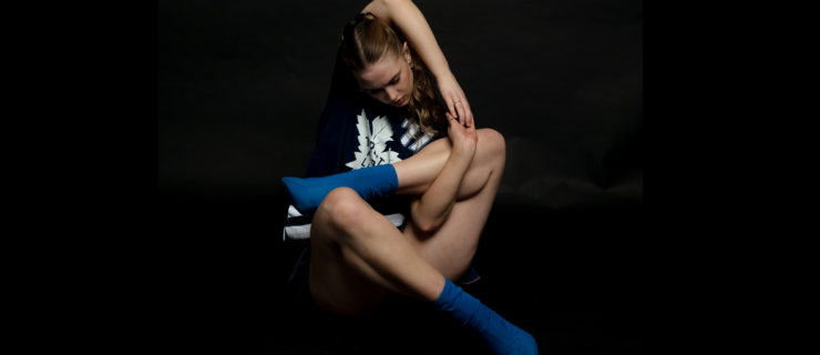 In front of a pitch-black backdrop, Lucy Turner sits with her limbs bent up and crossed in a pretzel-like shape, one arm reaching up and older to clasp her hands around one of her shins. She wears a black and white graphic t-shirt, black shorts, and blue socks.