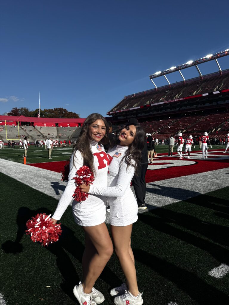Two dancers in Rutgers University Dance Team uniforms hug and pose for a photo on the football field. The dancer on the left holds a red pom out to the side.